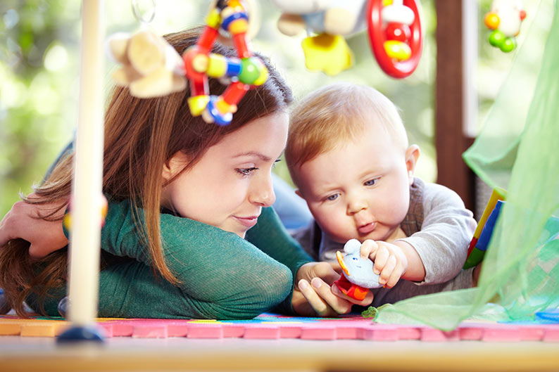 blonde baby boy playing with toys beside his mom