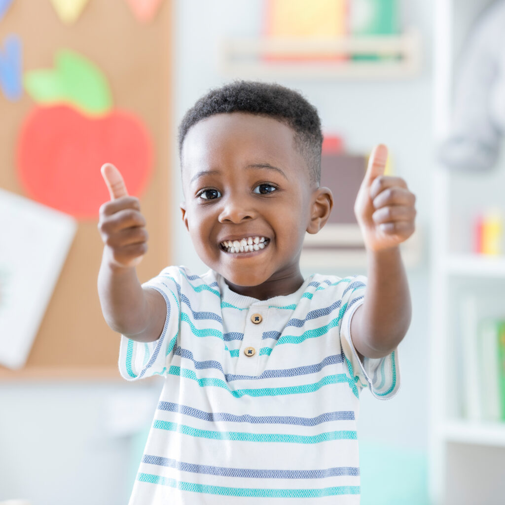 A young Black boy with a smile and thumbs up in a preschool classroom, radiating positivity and enthusiasm.