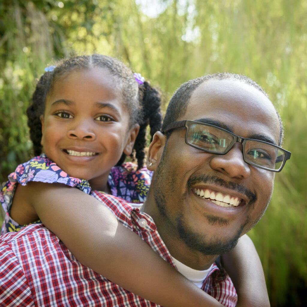 A little black girl on her dad's back, with her arms around him, both beaming with smiles towards the camera.