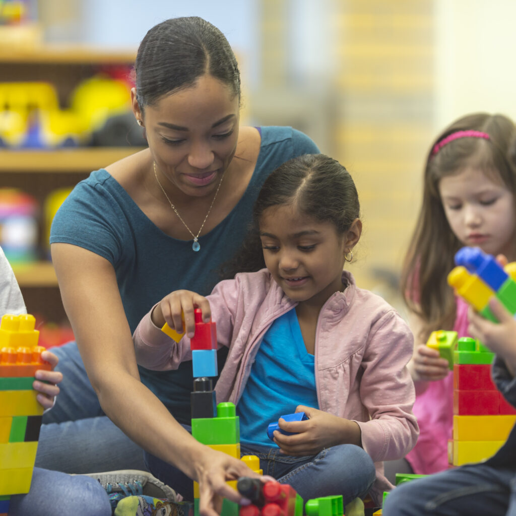A black caregiver engages in play with a small child, building legos together in a classroom setting.