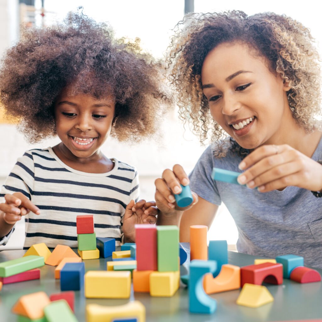 A black caregiver engages in play with a young black girl using building blocks.